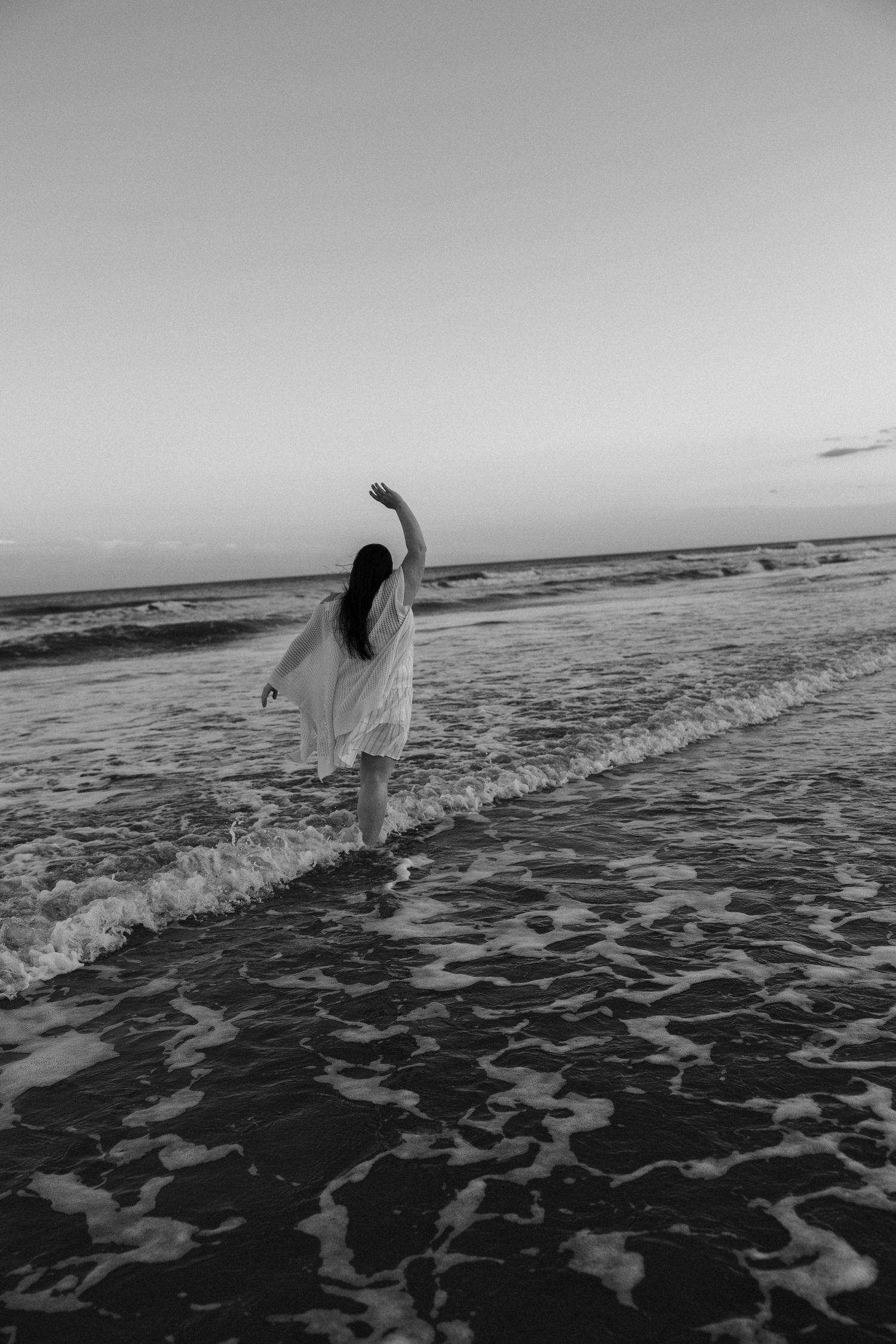 Person in white walking through ocean waves at sunset in black and white photography.