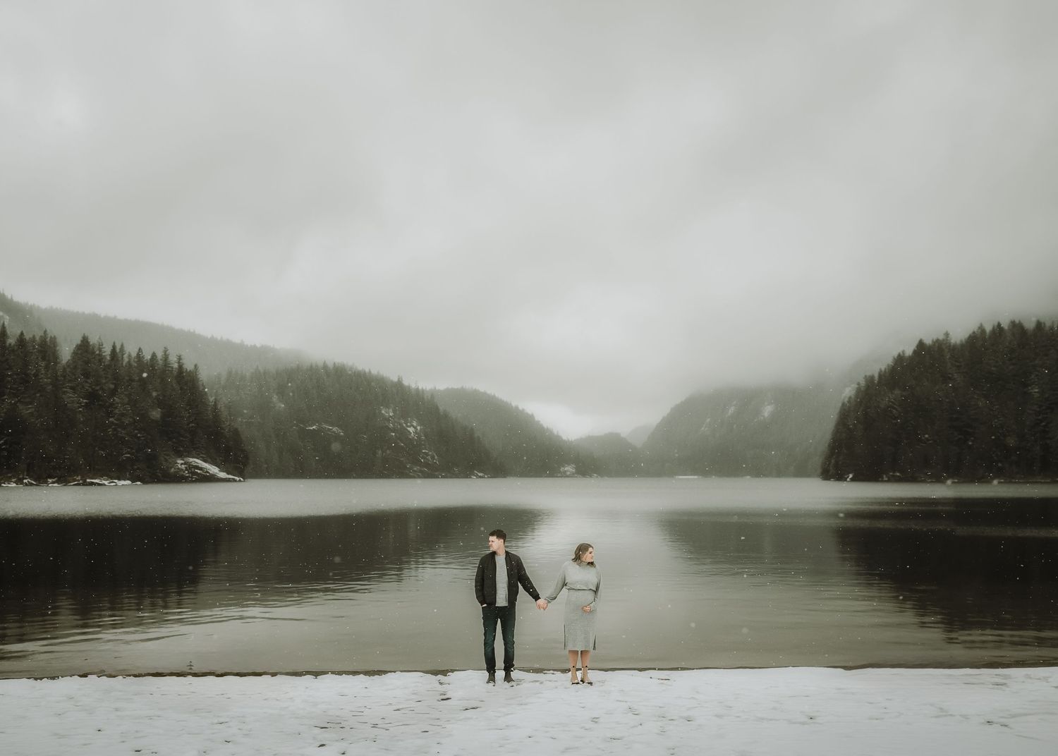 A couple stands at the edge of a misty lake surrounded by evergreen forests and mountains in moody winter scene.
