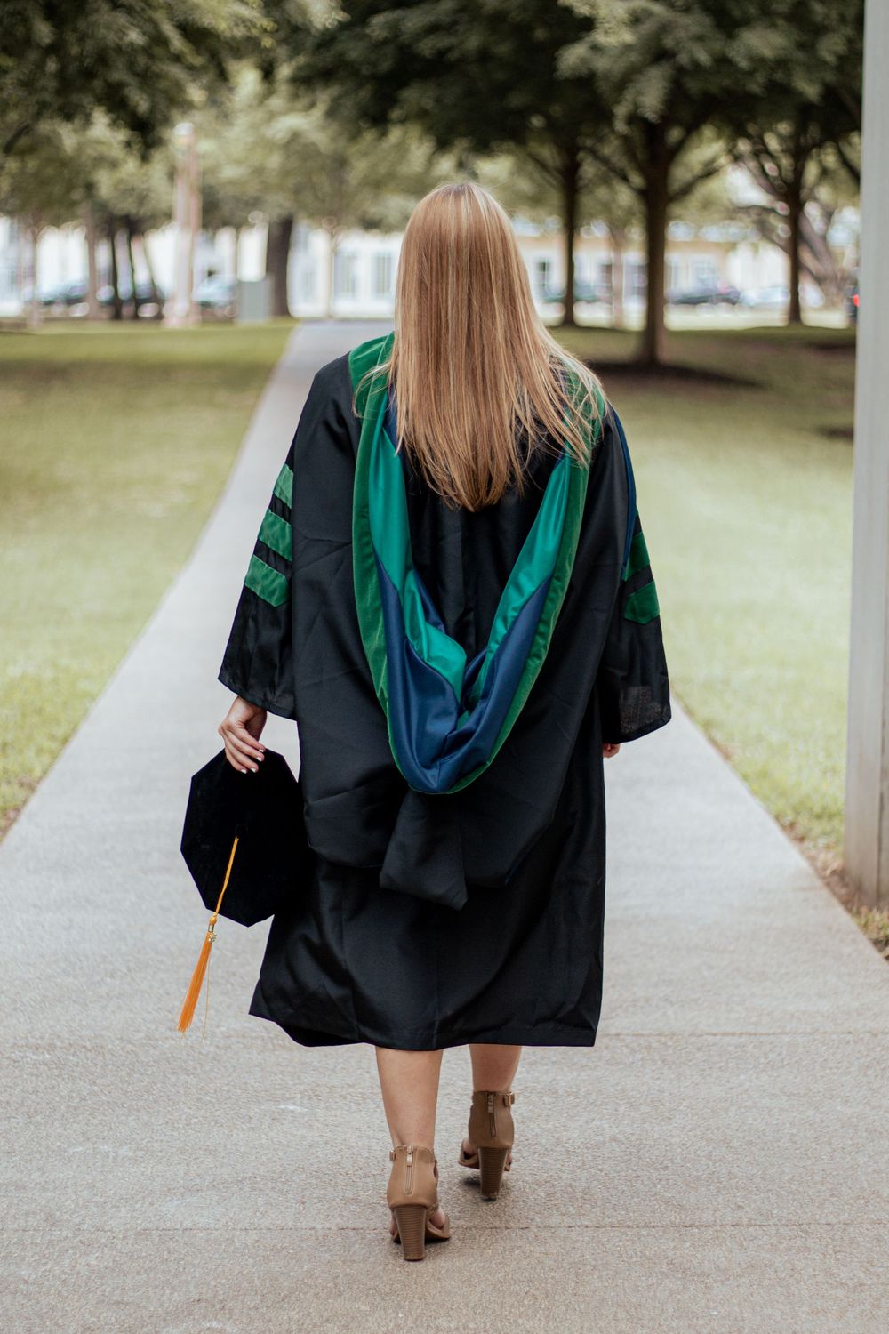 Graduate in black cap and gown with green hood walking away on campus sidewalk carrying diploma.