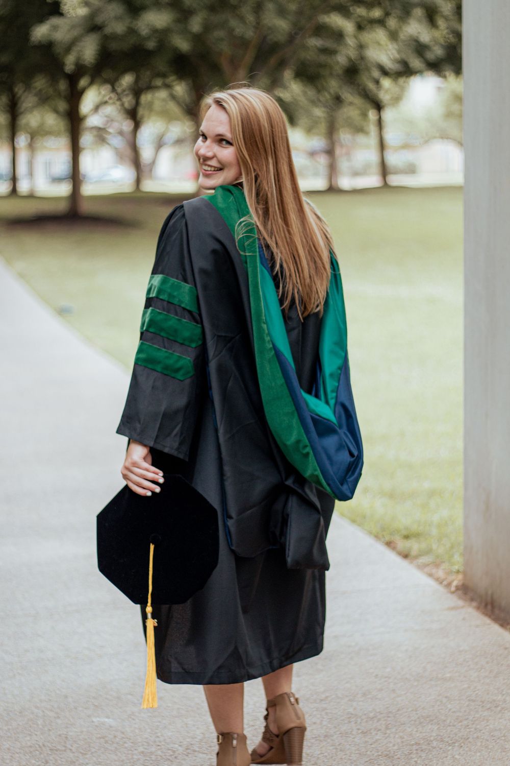 Graduate wearing traditional black academic regalia with green stripes and tassel stands on pathway near trees.