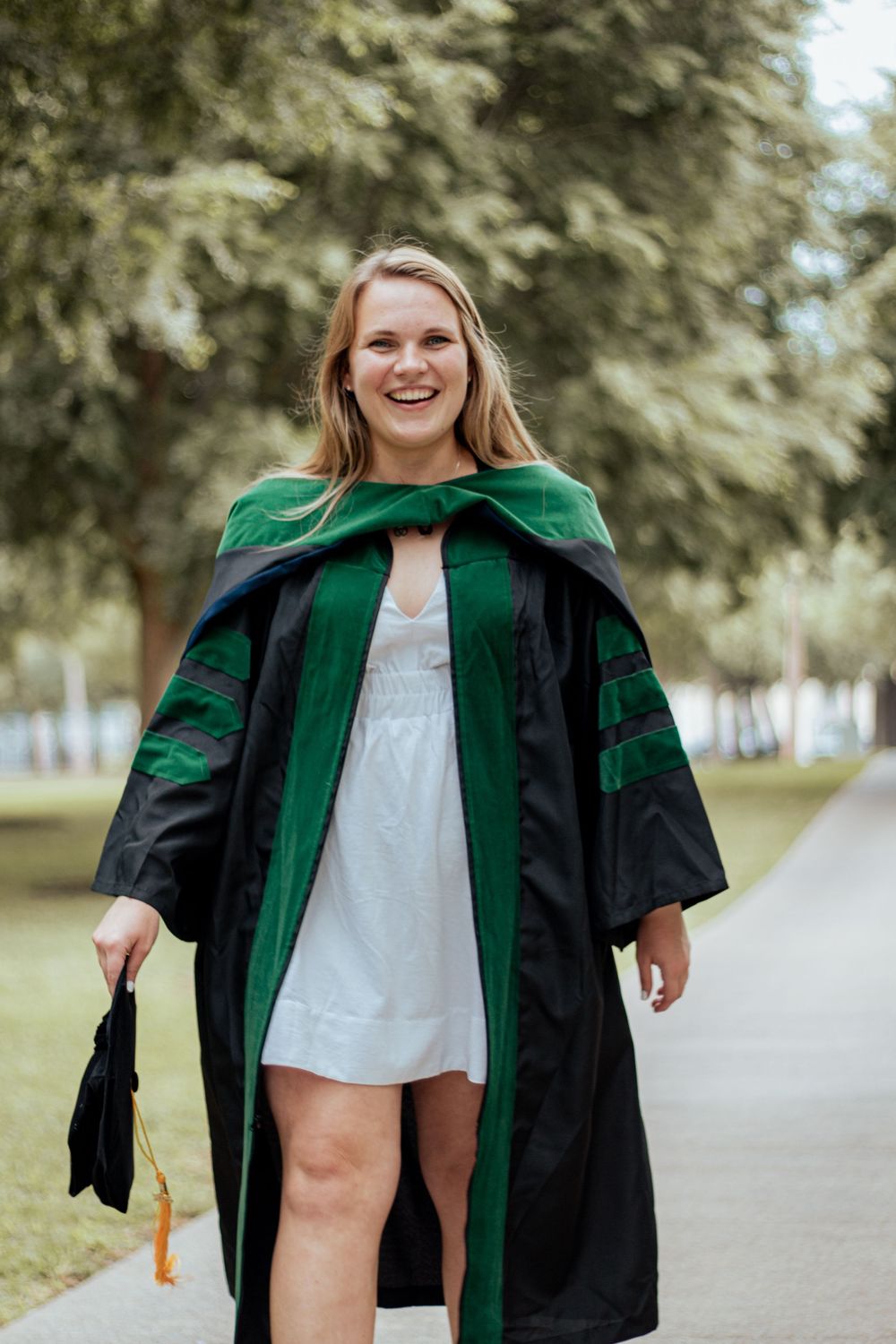 Graduate wearing traditional black and green academic regalia walks down campus pathway smiling.