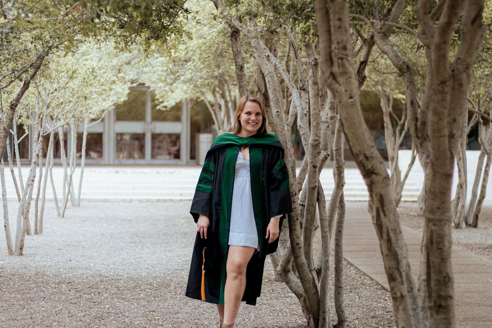 Graduate in black gown with green hood walks along tree-lined path during graduation photoshoot on campus.