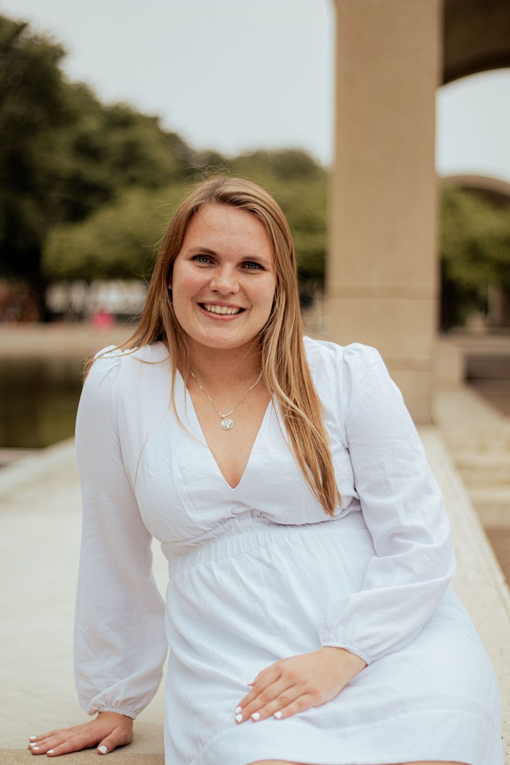 Professional portrait of a woman in a white dress with long sleeves sitting outdoors by a column.