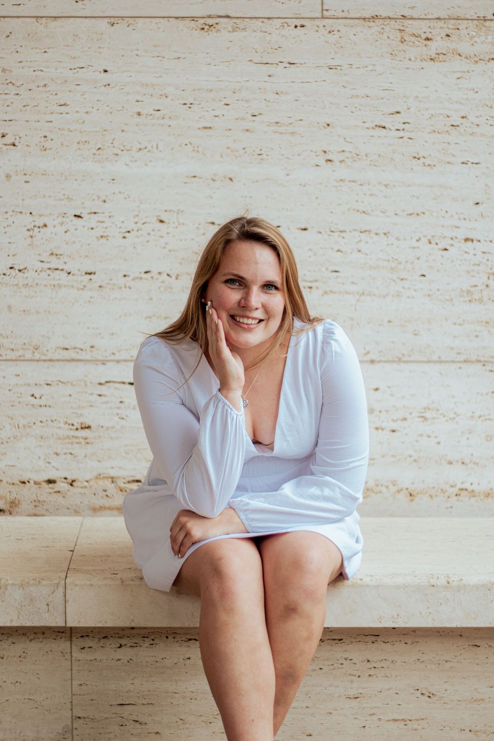 A person in a white outfit sits casually against a textured beige wall while resting their hand on their chin.