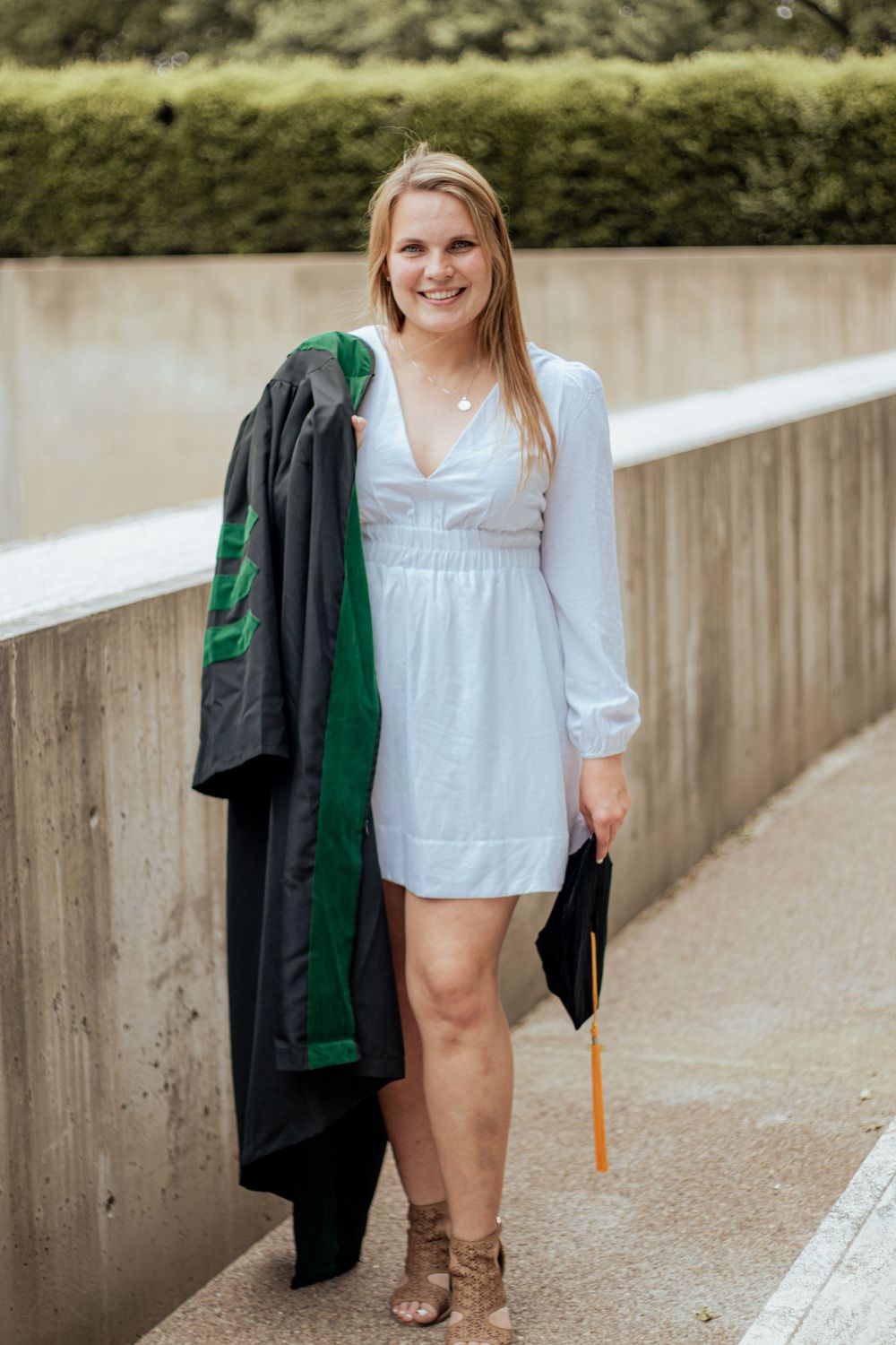 Someone in a white long-sleeved dress holds a black and green coat while standing along a concrete path outdoors.