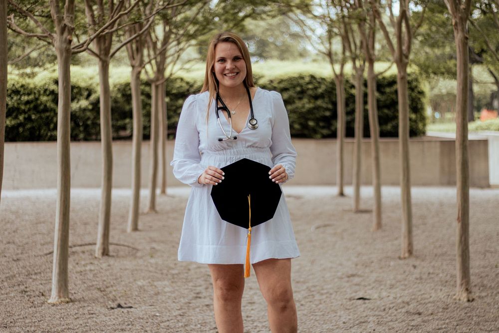 A person in a white dress smiles while standing in a path lined with evenly spaced trees.