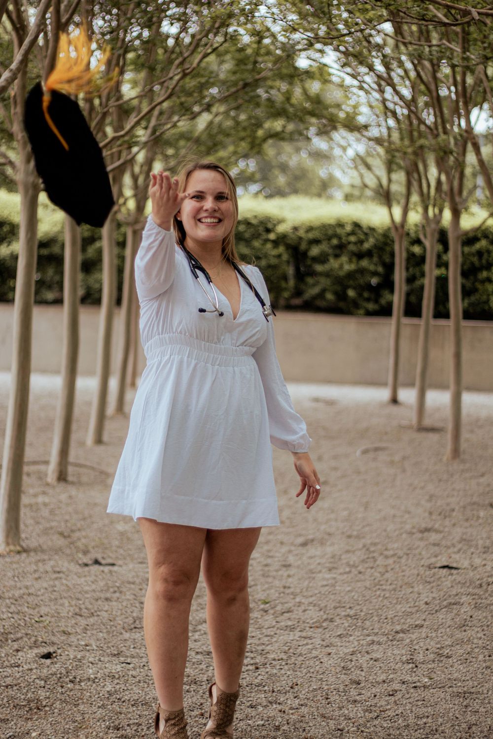 A person in a white summer dress and cowboy boots stands on a dirt path surrounded by trees.