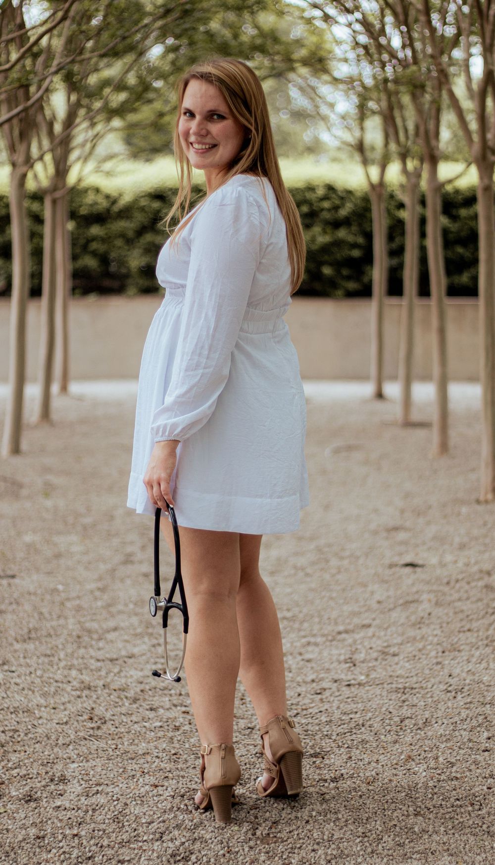 A person in a white summer dress standing in an outdoor garden setting lined with tall trees.
