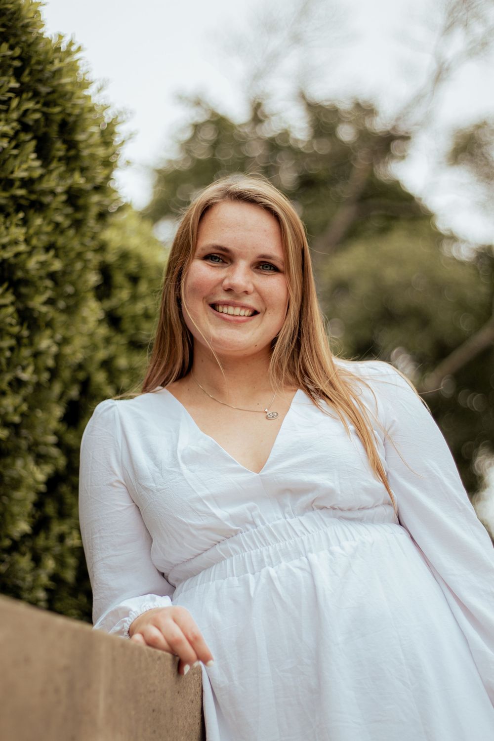 Portrait of someone in a white dress smiling outdoors with greenery in the background on a sunny day.