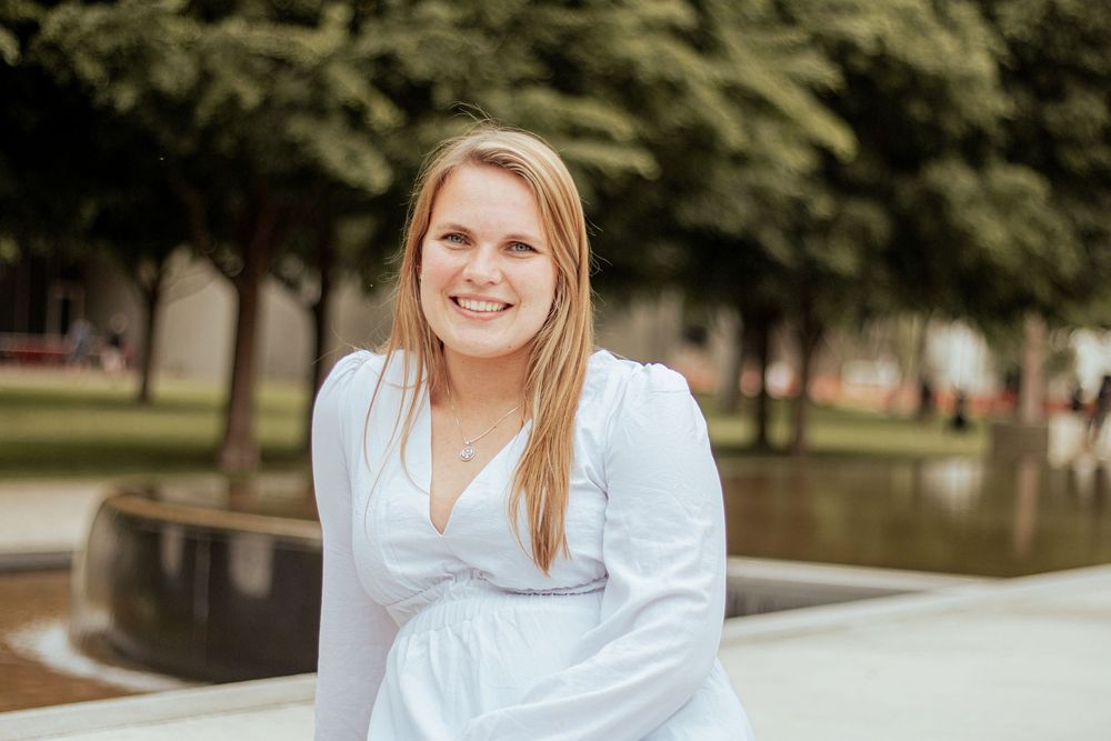Person in white dress sitting on edge of outdoor fountain in park with trees in background.