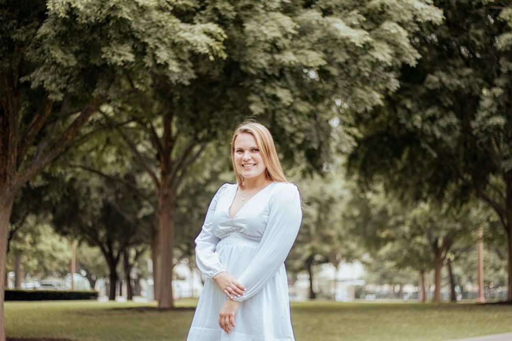 Expectant woman in a light blue dress stands in a serene park setting surrounded by green trees.
