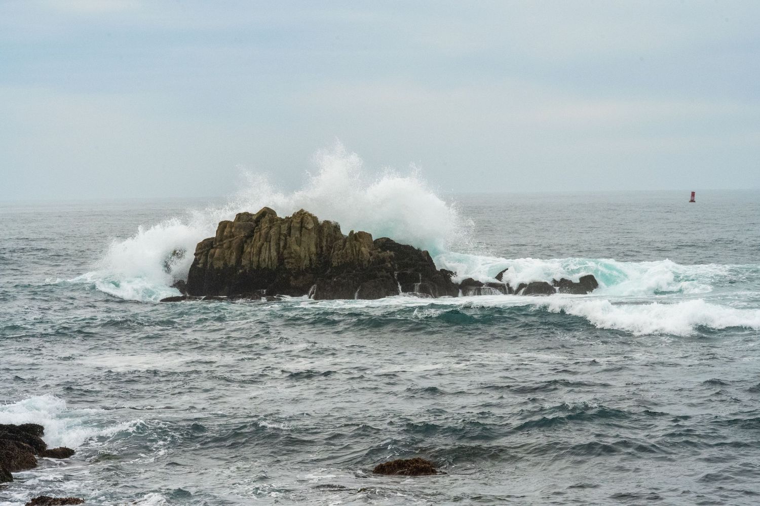 Powerful waves crash against a rocky outcrop in a stormy ocean with gray skies overhead.