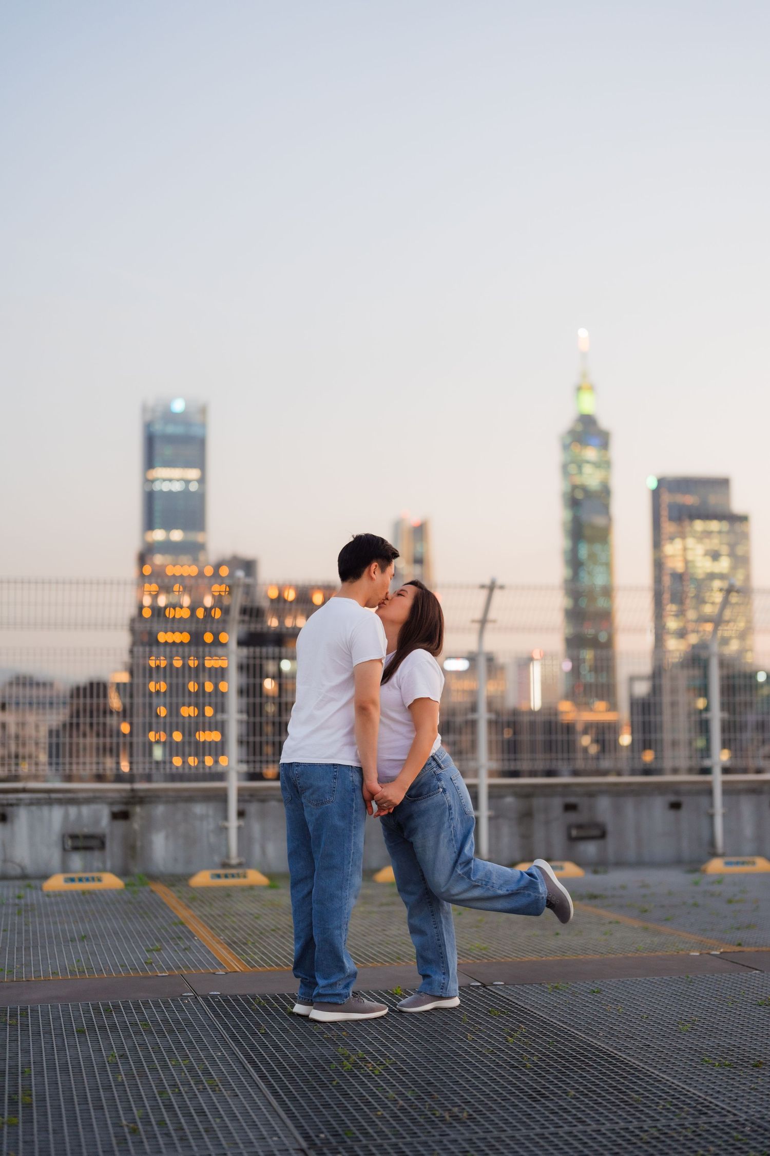 A couple shares a romantic moment on a rooftop parking deck with Taipei 101 and the city skyline glowing at dusk.