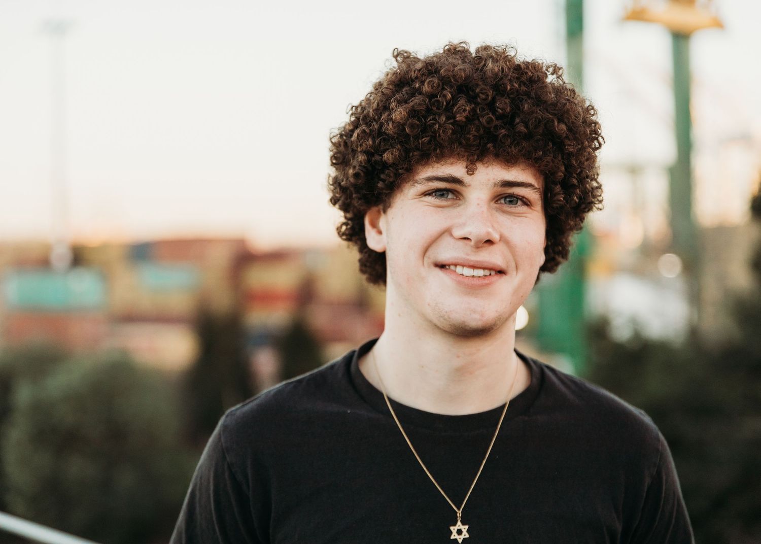 Portrait of a young person with curly dark hair and a necklace smiling outdoors at sunset.