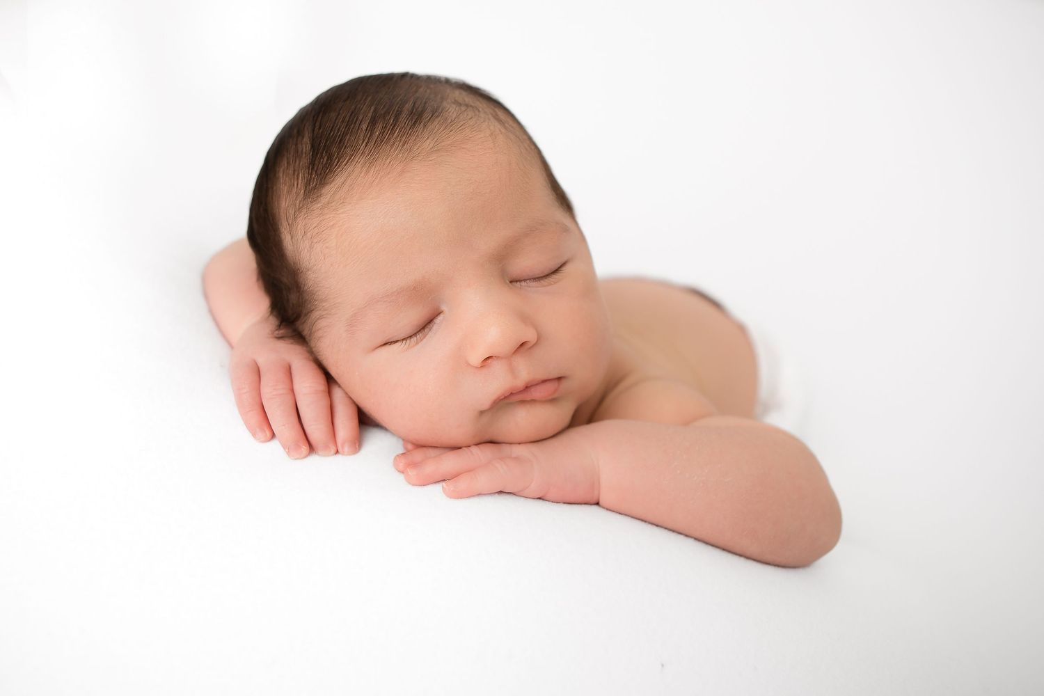 A peaceful sleeping newborn baby rests with head on folded arms against a white background.