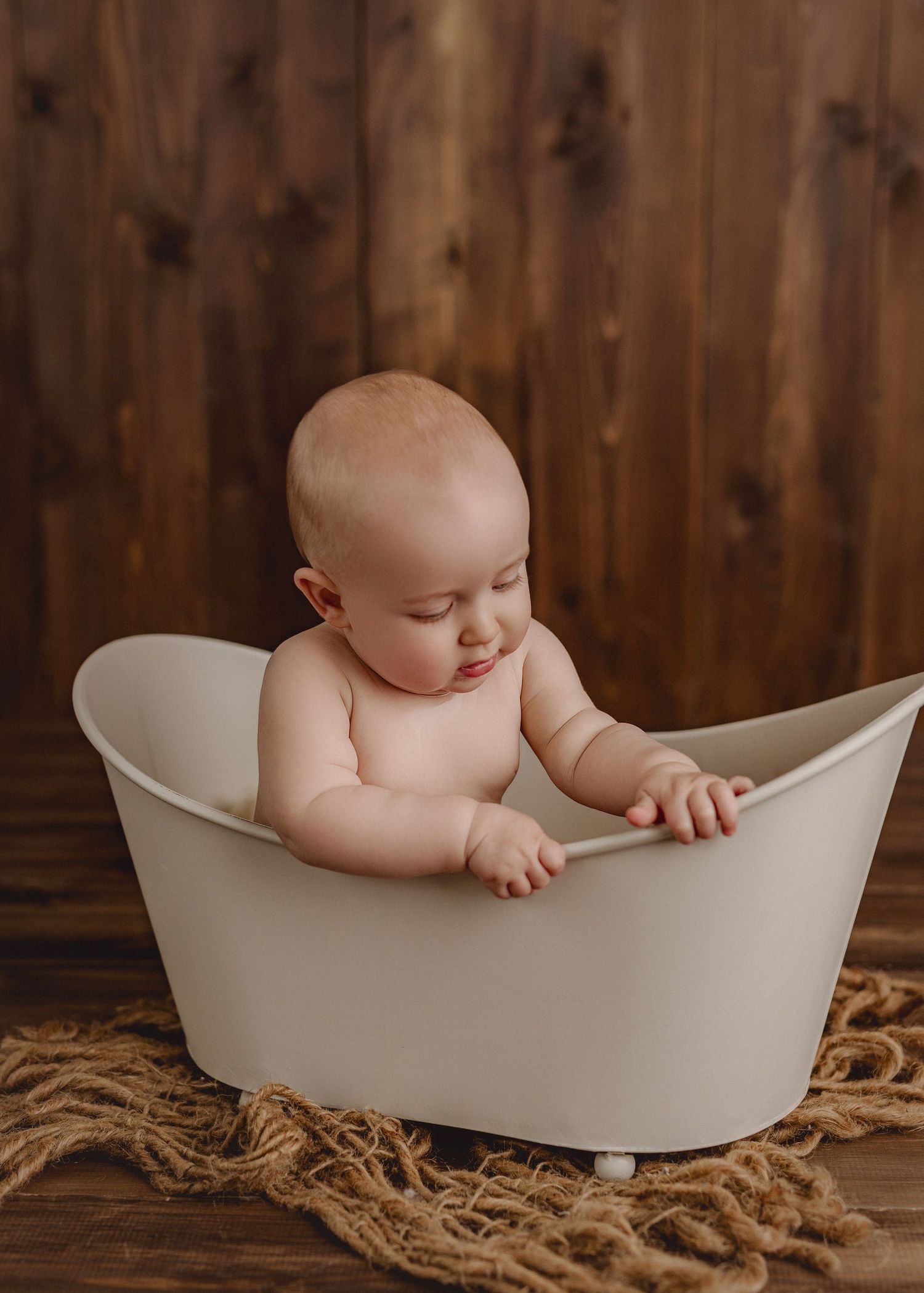 Sweet baby sits in a white modern bathtub with a rustic wooden wall background and jute rug underneath.