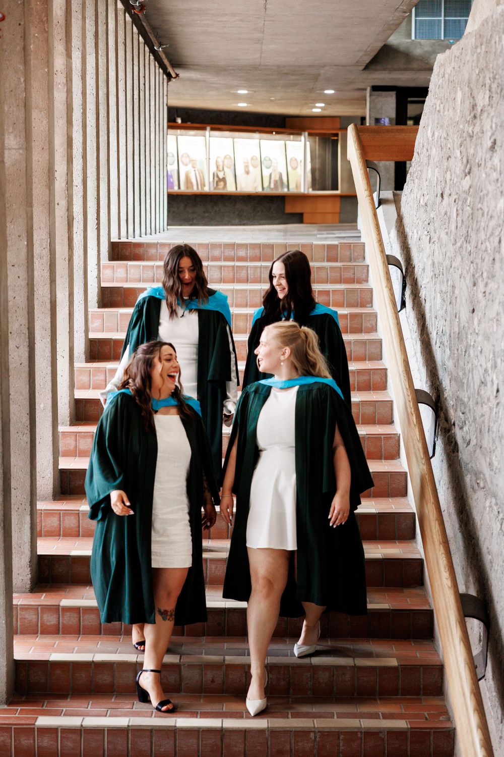 Students in matching green graduation robes and white dresses walking down brick stairs in a school building interior.