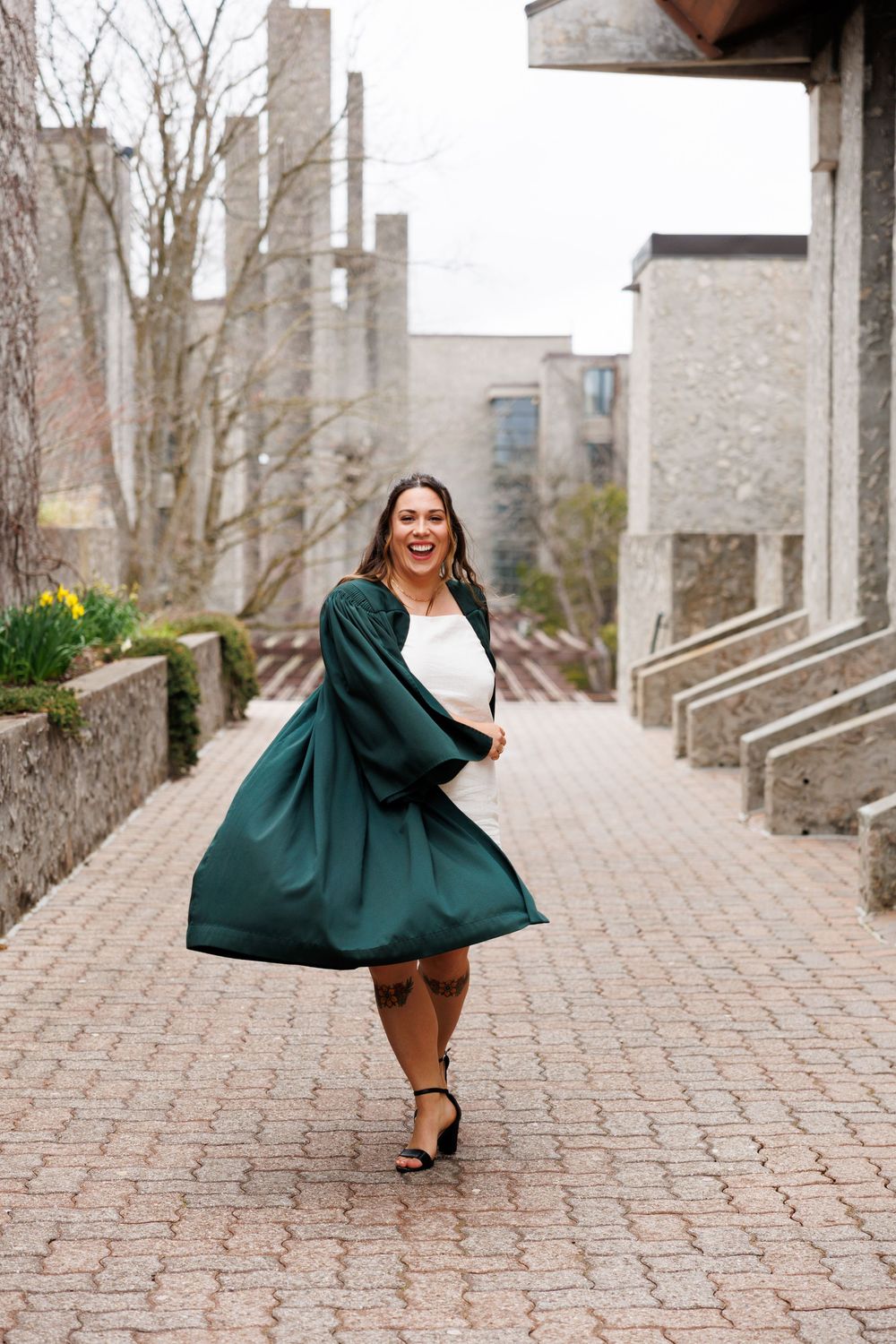 Someone dances joyfully on a brick path wearing a flowing emerald green graduation gown and white dress underneath.