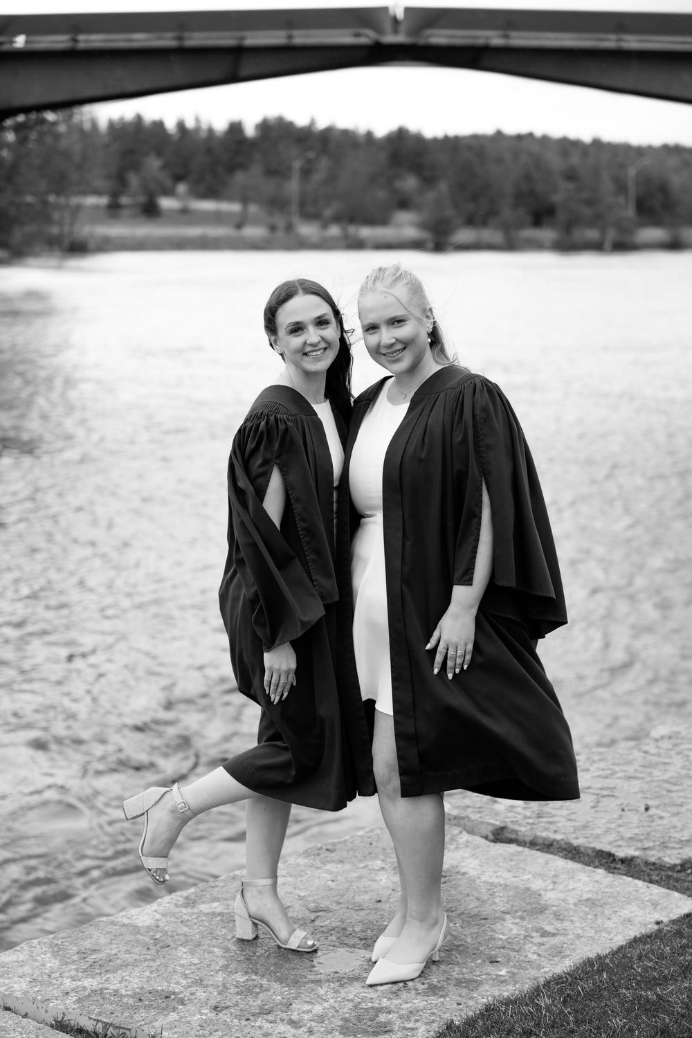 Two graduates in black academic gowns pose together near a lake for a graduation photo shoot in black and white.