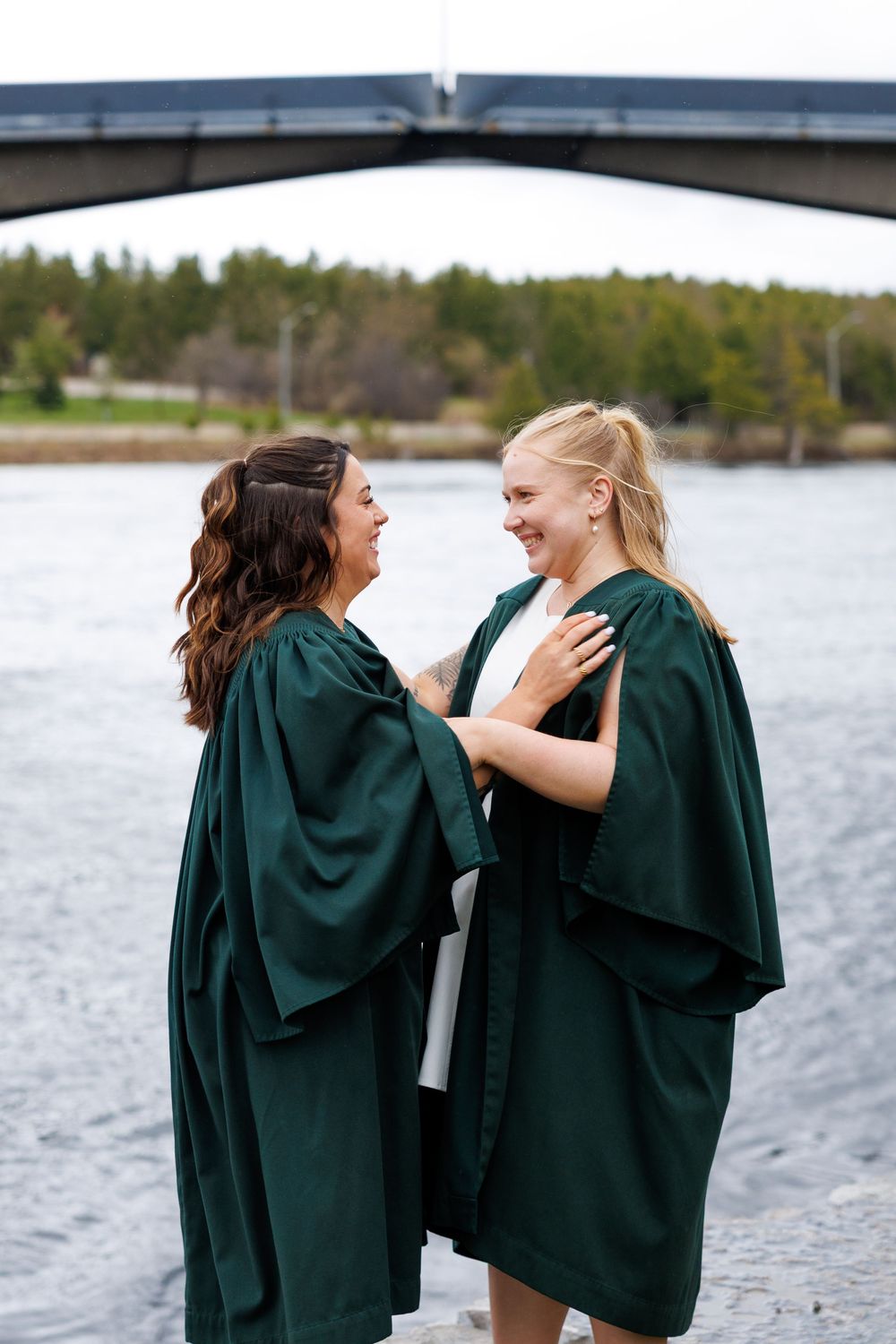 Two graduates in dark green graduation gowns share a joyful moment by a lake during commencement celebrations.