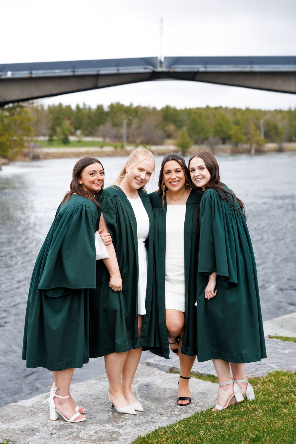 Four graduates in dark green academic robes pose together by a lake with a bridge in the background.
