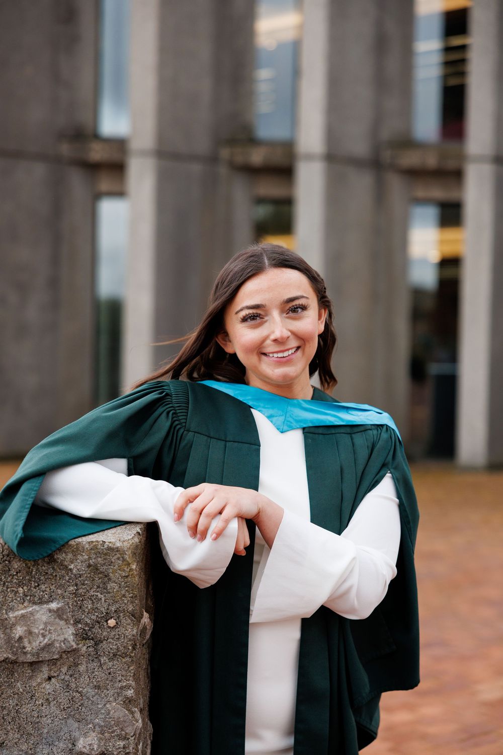 A graduate in green academic regalia and white sweater poses outside a building with stone columns.