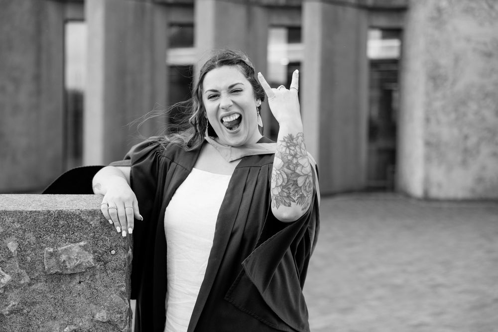 A candid black and white portrait showing an exuberant expression and visible arm tattoo against an urban backdrop.