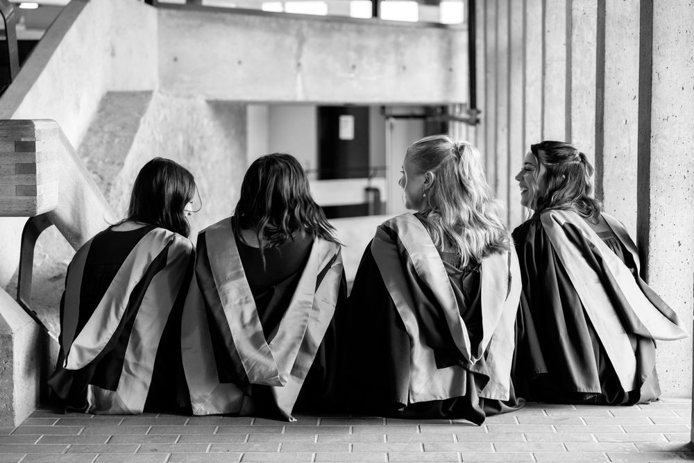 Four students wearing graduation gowns sit together on steps in a black and white photograph.