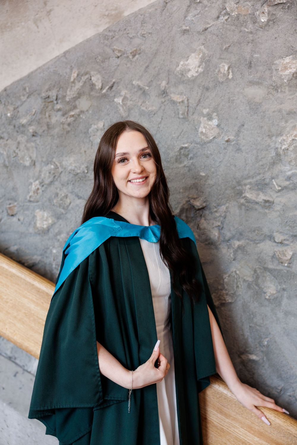Graduate wearing academic regalia including dark green graduation gown and blue stole poses against textured wall.