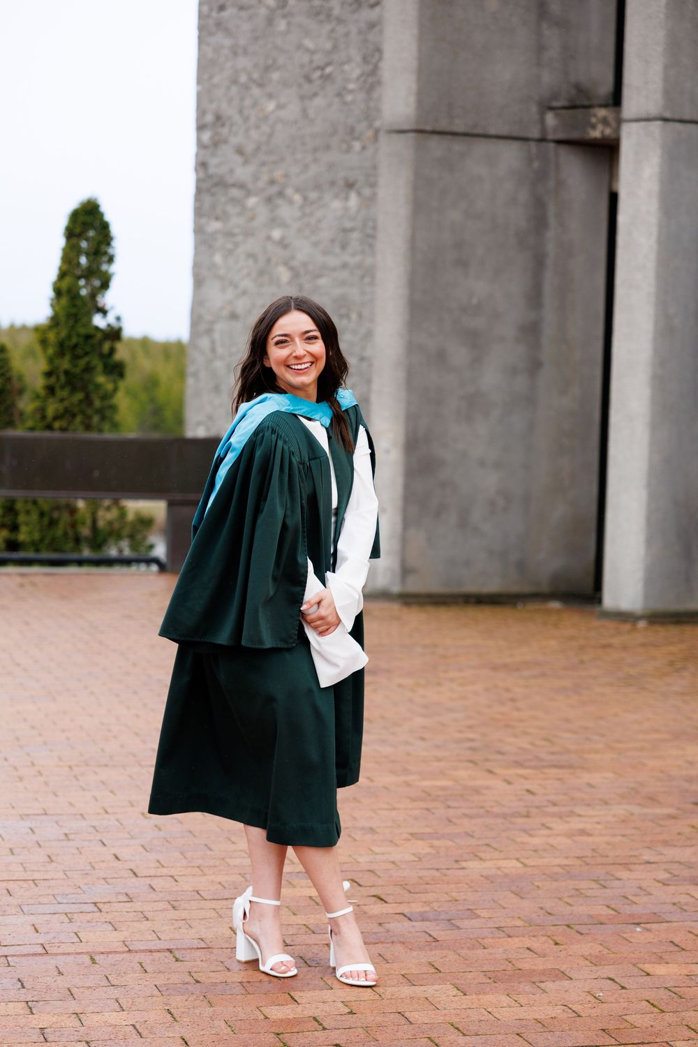 Graduate in an emerald green academic gown and blue stole poses smiling on brick pavement near concrete pillars.