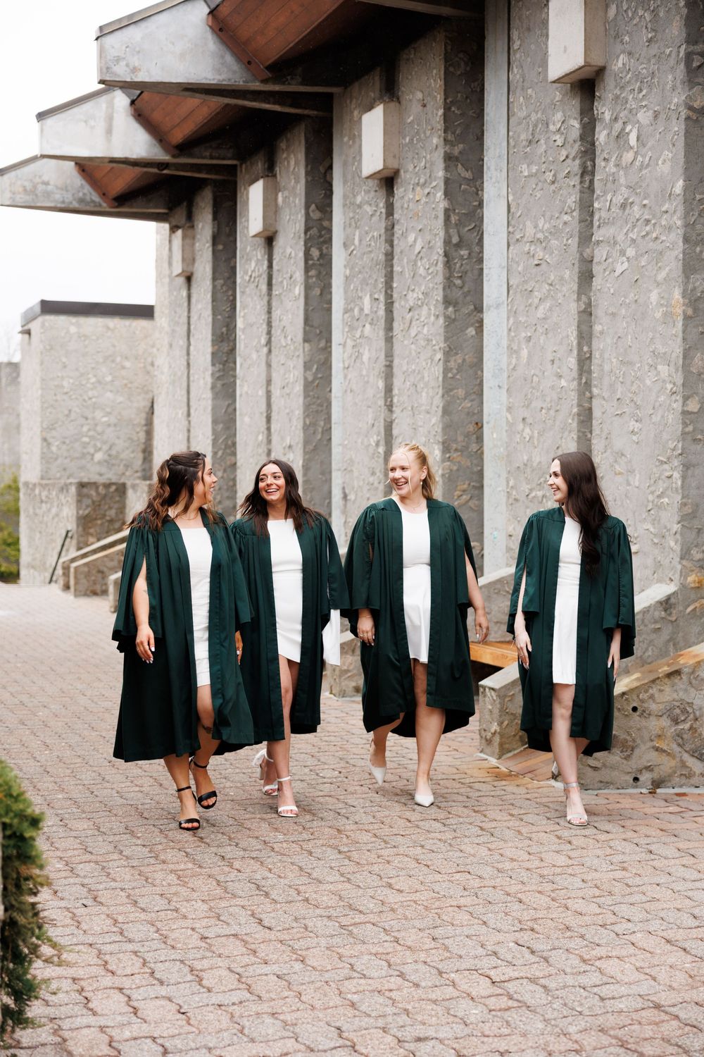 Four graduates wearing dark green graduation gowns walk together on a stone pathway outside a modern building.
