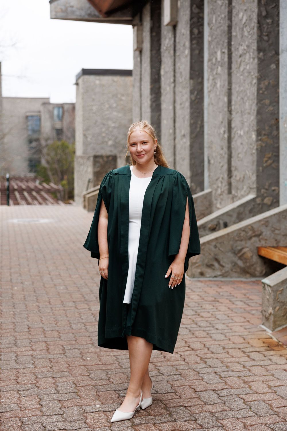 Graduate wearing dark green academic regalia gown and white dress stands on brick pathway near stone columns.