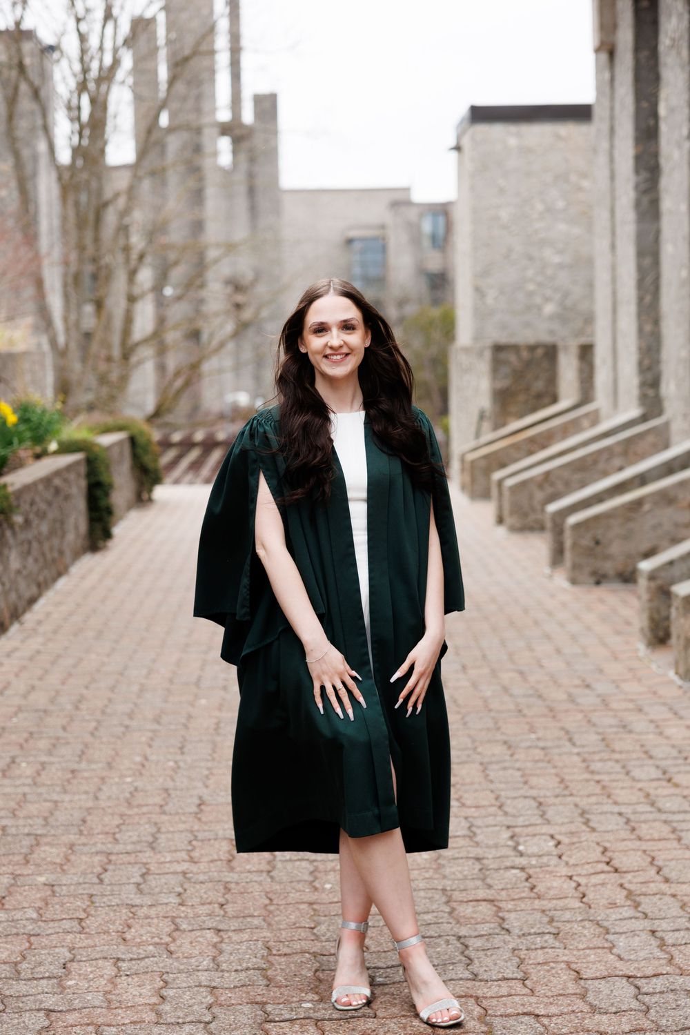 Graduate in dark green academic regalia poses on brick pathway by stone university building.