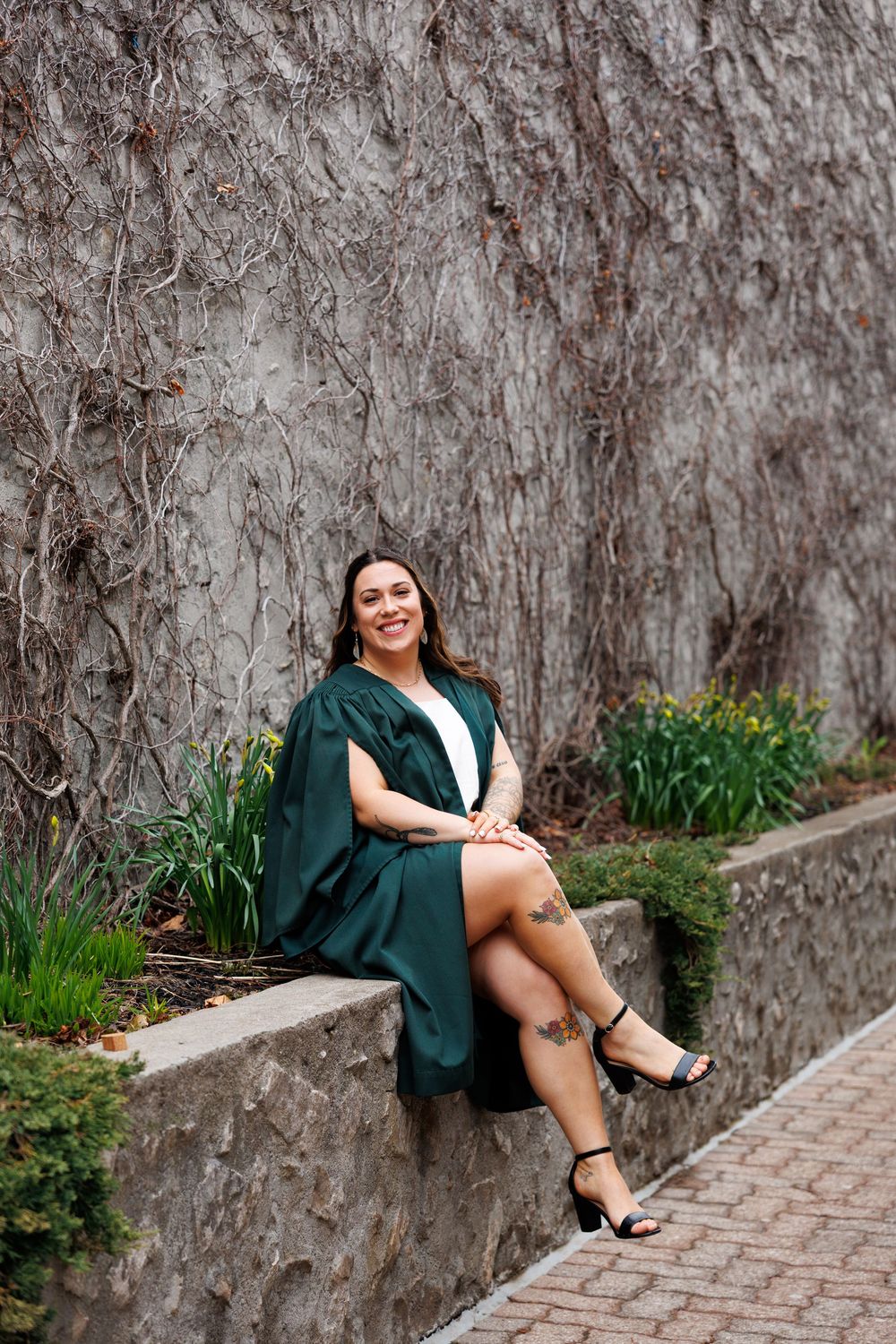 Graduate in green cap and gown sits casually on stone wall with ivy and spring flowers blooming in background.