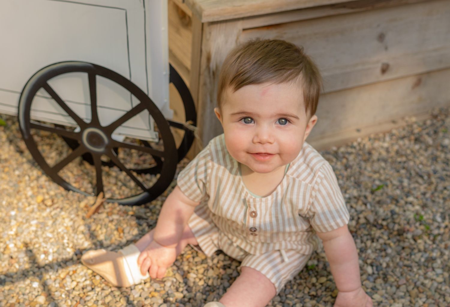 Baby in striped outfit sitting outdoors on gravel surface.