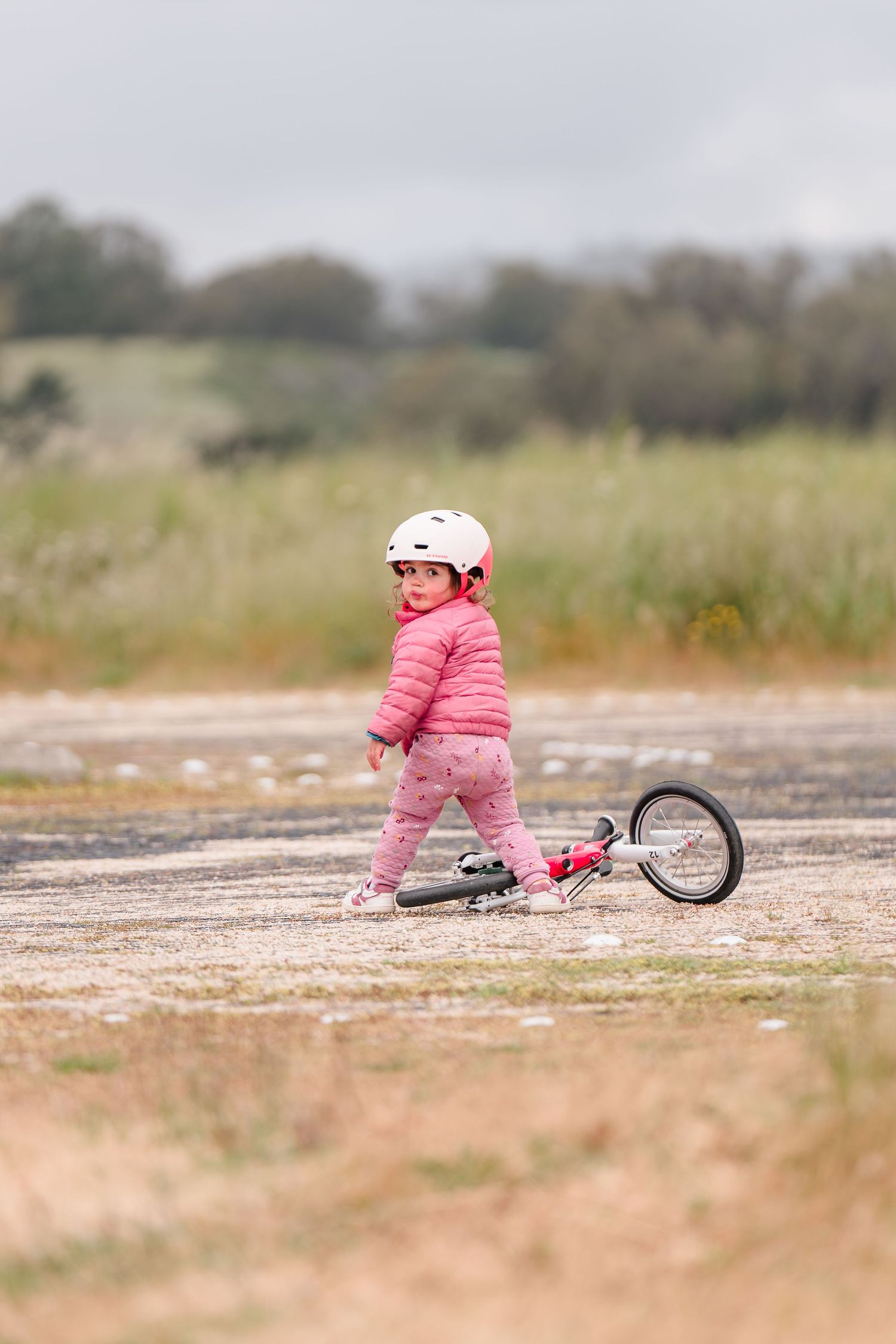 A pink-clad child falls over while learning to ride a bicycle on a dirt path. Located in UCSC Seymour marine center in Santa Cruz West side.