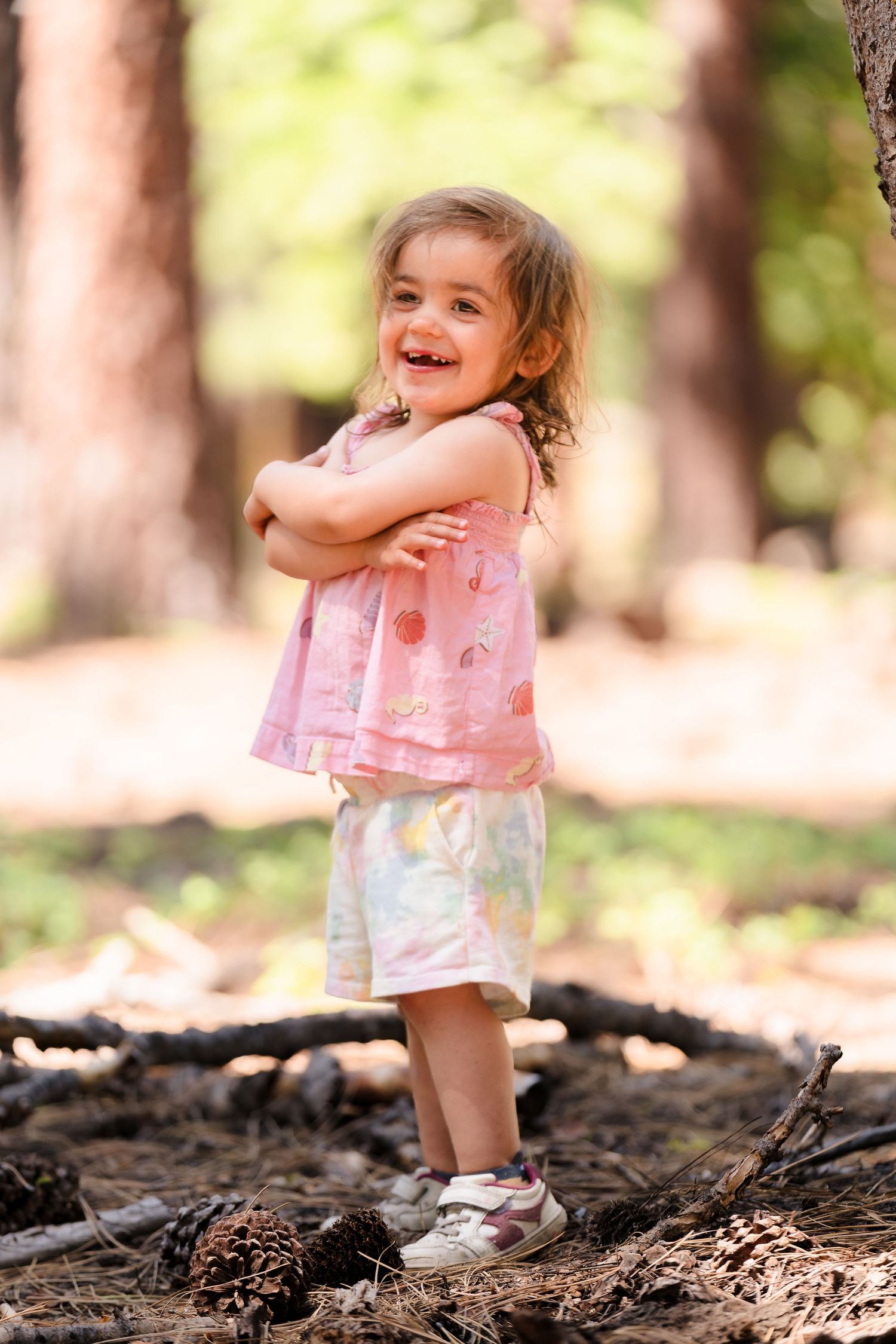 Young child in a pink top and tie-dye shorts stands playfully on a forest trail with hands crossed and a bright smile. Located in Felton.
