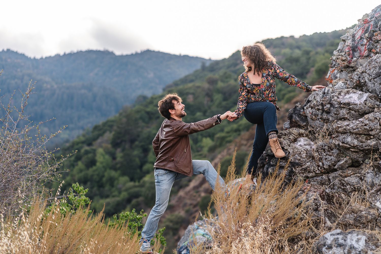 Two people share a happy moment while hiking on rocky terrain with mountains in the background at sunset. Located near Felton.