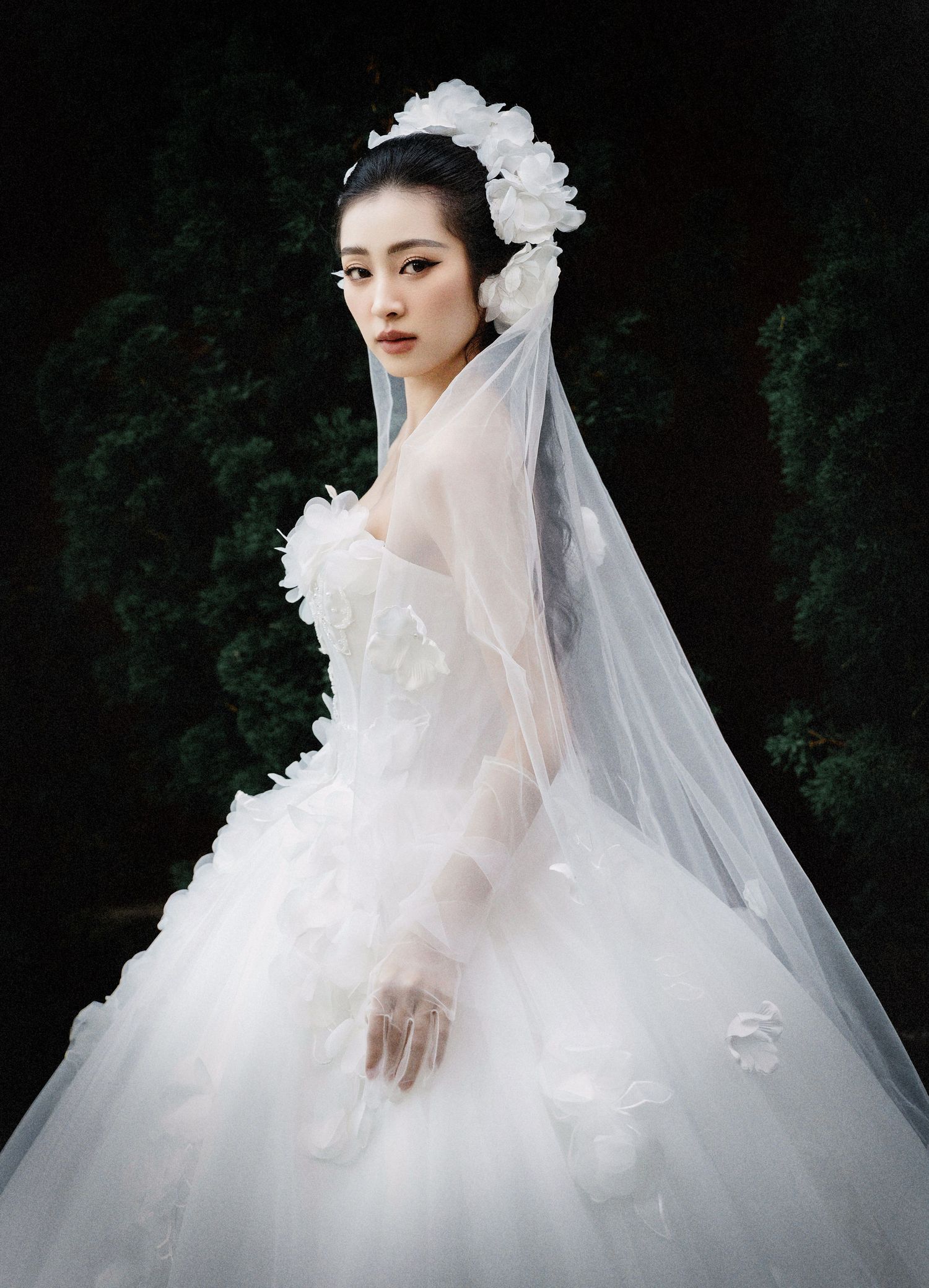 A bride in a white ball gown with sheer gloves and floral headpiece stands elegantly against a garden backdrop with statue.