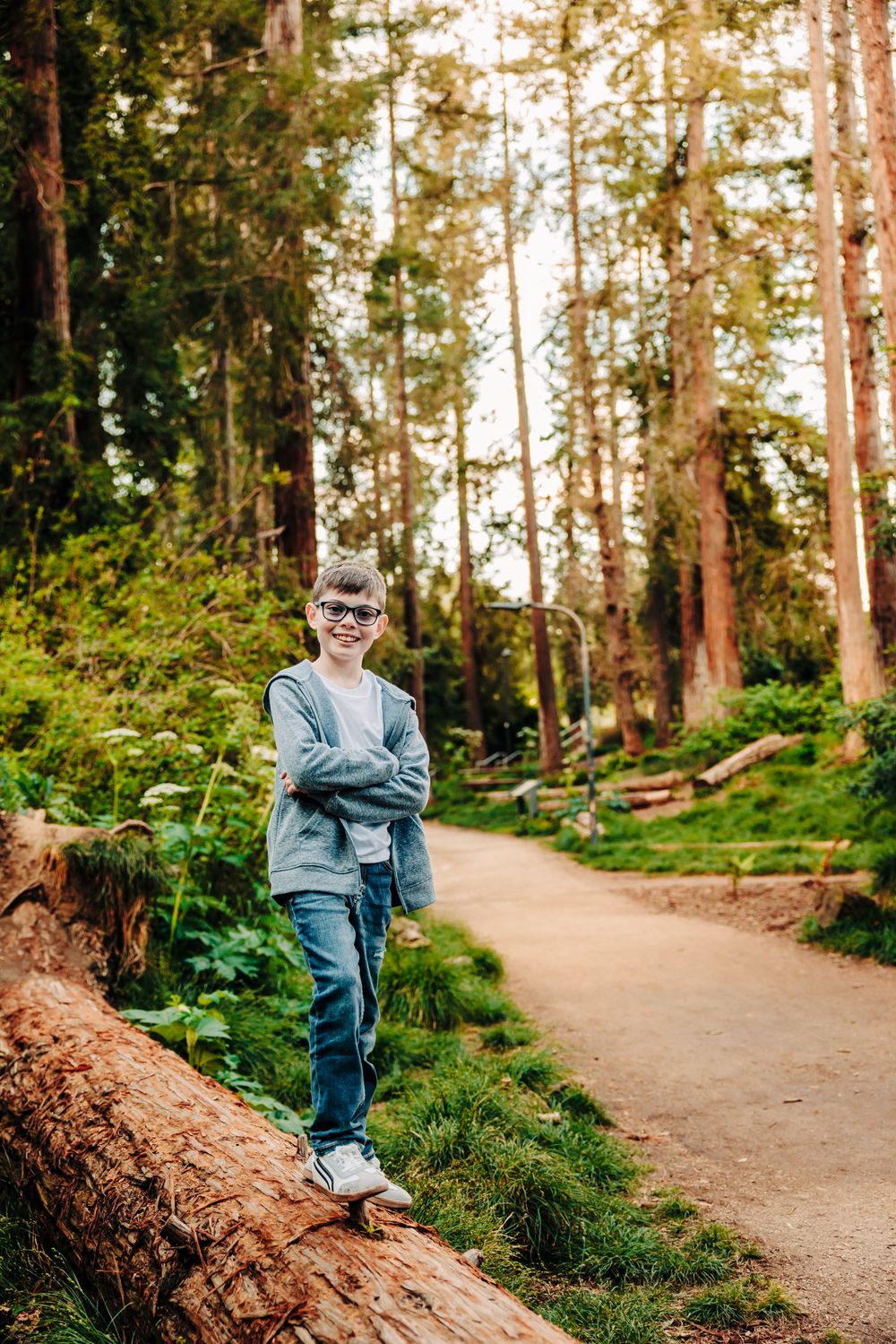 Child sitting on fallen log along a winding forest trail surrounded by towering redwood trees.