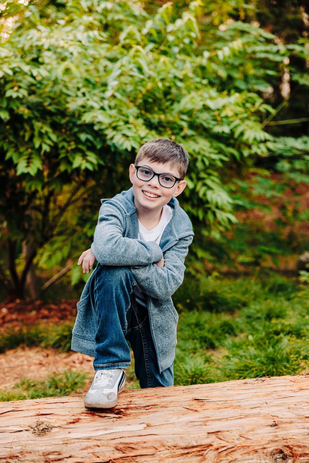 A young child sits casually on a large fallen log in a lush green forest setting.