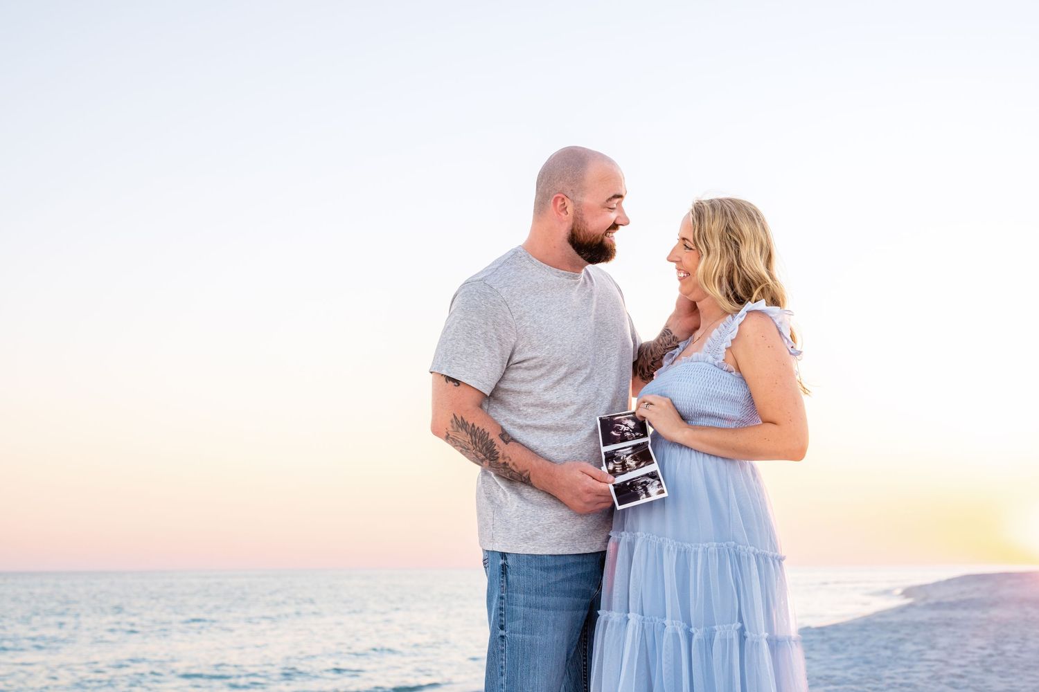 A couple stands together at the beach during sunset, smiling and holding ultrasound photos.