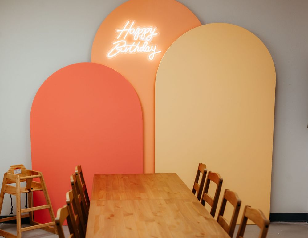 Modern restaurant dining area with wooden table and chairs against curved coral and beige wall panels with neon signage.