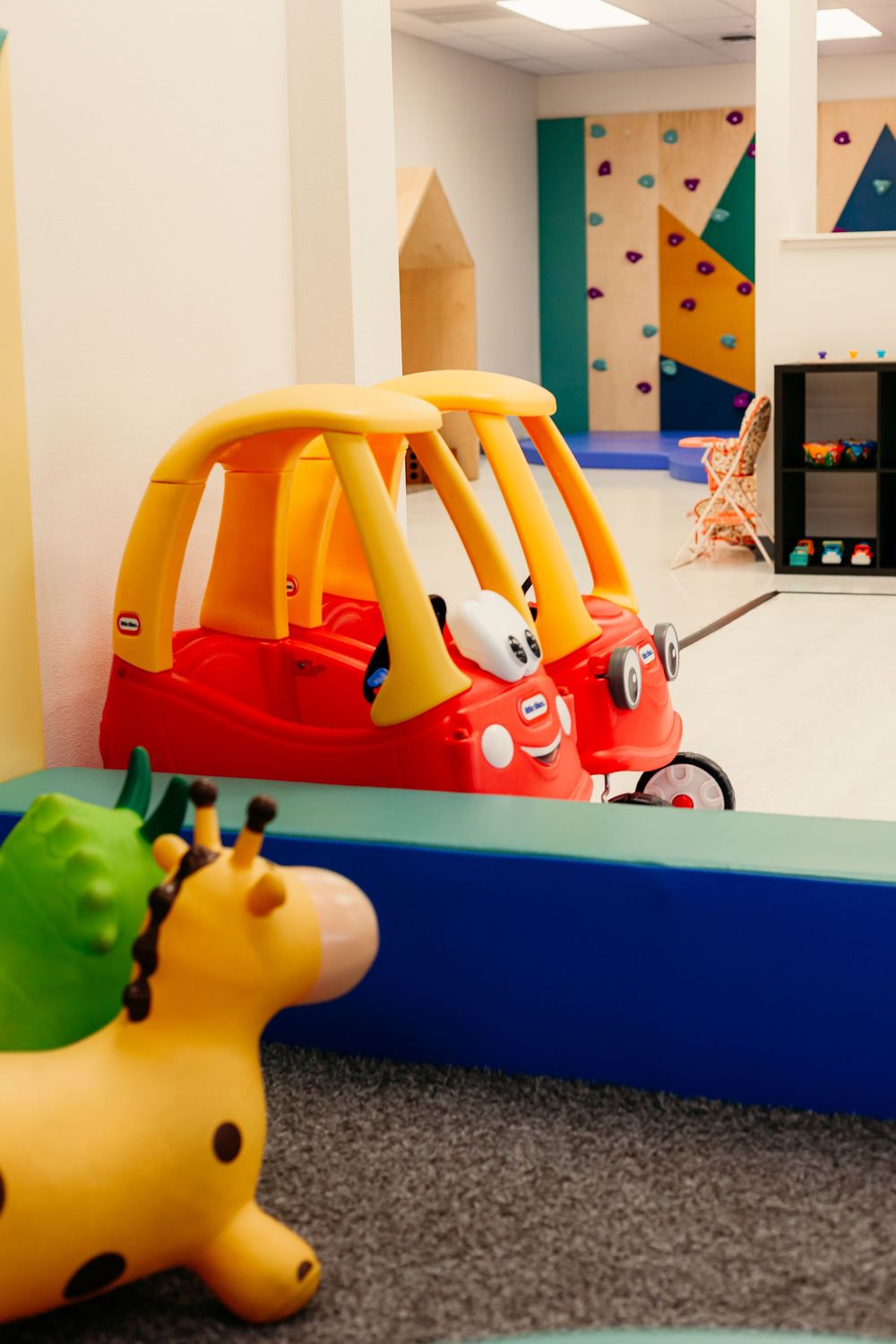 Red and yellow toy cozy coupe car in indoor play area with climbing wall background and rubber giraffe toy.