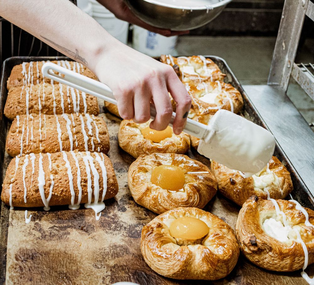 Freshly baked Danish pastries with icing drizzle and custard filling being arranged on a baking sheet.