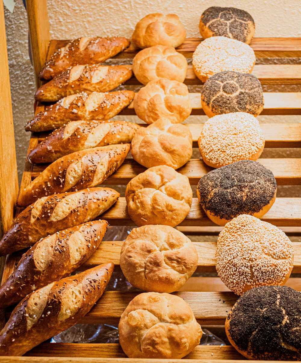 Fresh artisanal breads including twisted pretzel rolls, plain rolls, and seeded buns arranged on wooden bakery shelves.