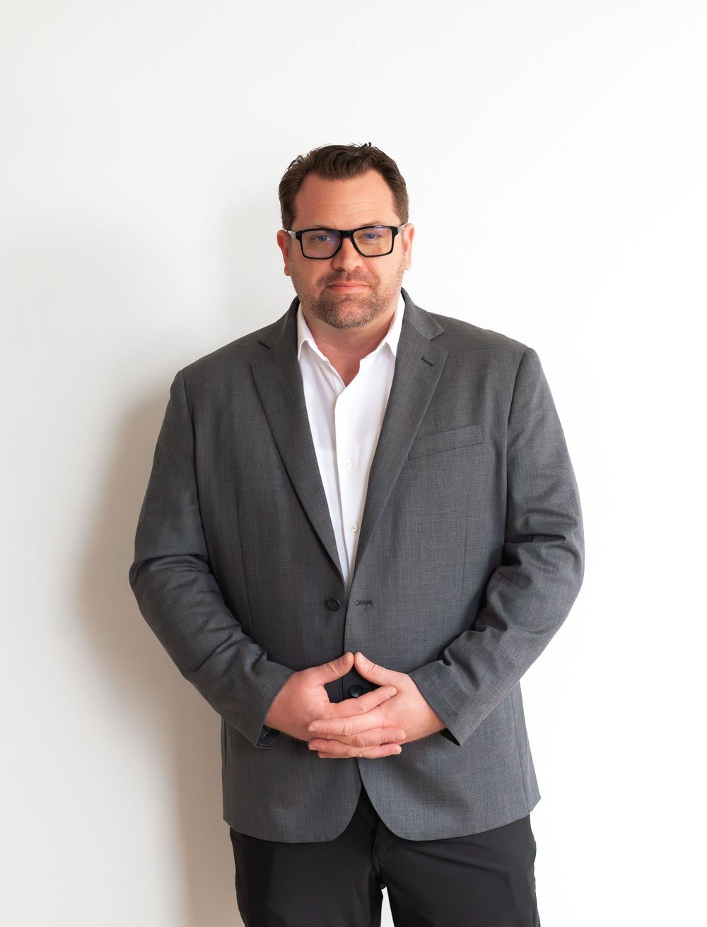 Professional wearing a grey suit jacket and white button-down shirt stands against white wall with clasped hands.