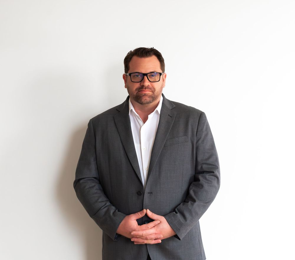 Professional headshot of a person in a dark gray suit and white shirt standing against a white wall.
