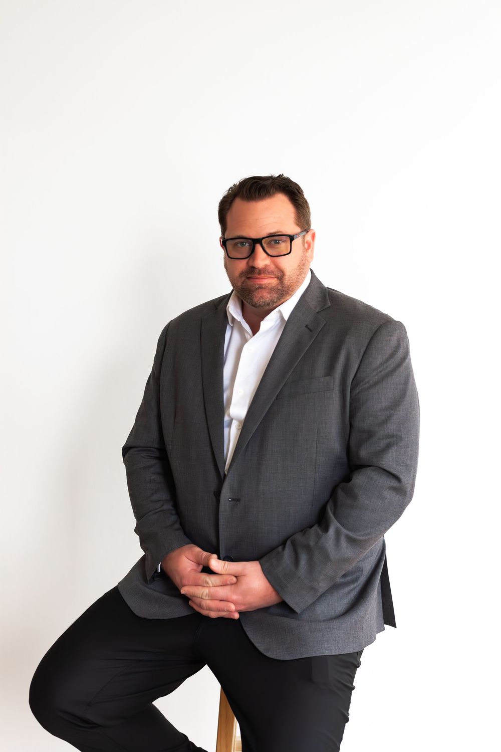 Professional studio portrait of someone in a gray suit jacket and glasses seated against a white background.