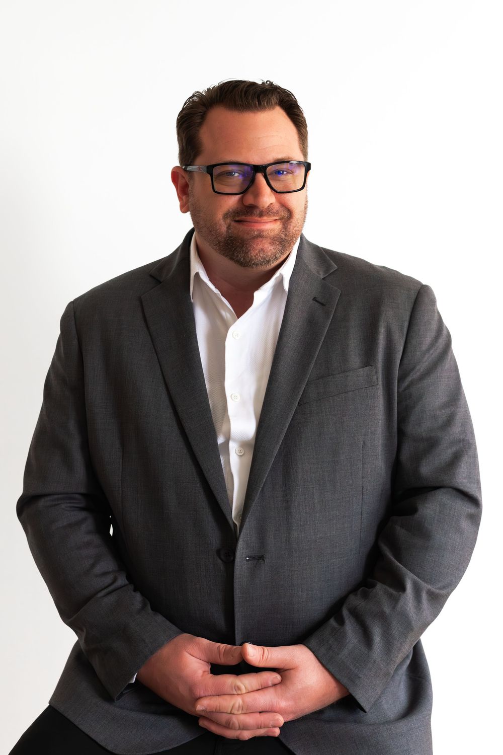 Professional headshot of a smiling person wearing glasses, a white dress shirt and grey suit jacket against light background.